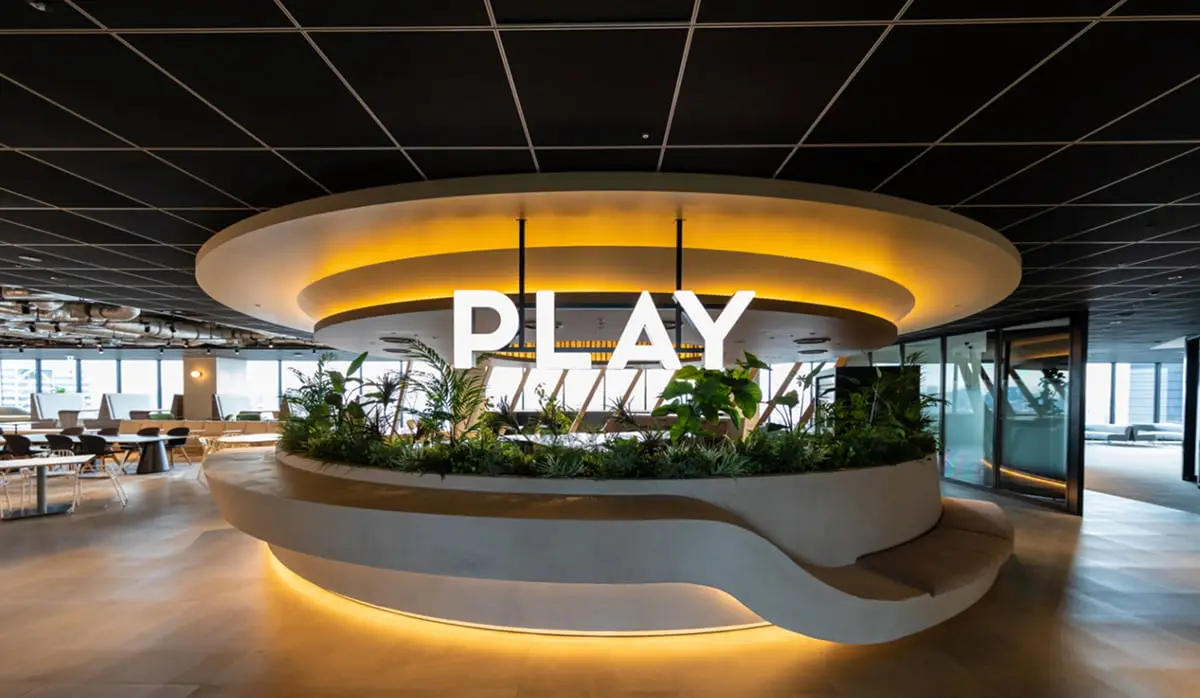 モダンなオフィスの中央に、植物が植えられた円形のベンチと「PLAY」の文字が配置された、明るく開放的な空間