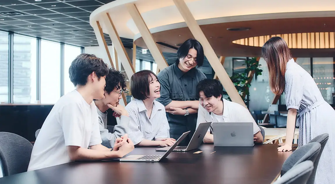 モダンなオフィスで、6人の若いプロフェッショナルが大きなテーブルを囲んでノートパソコンを使いながら活発に共同作業をしている様子