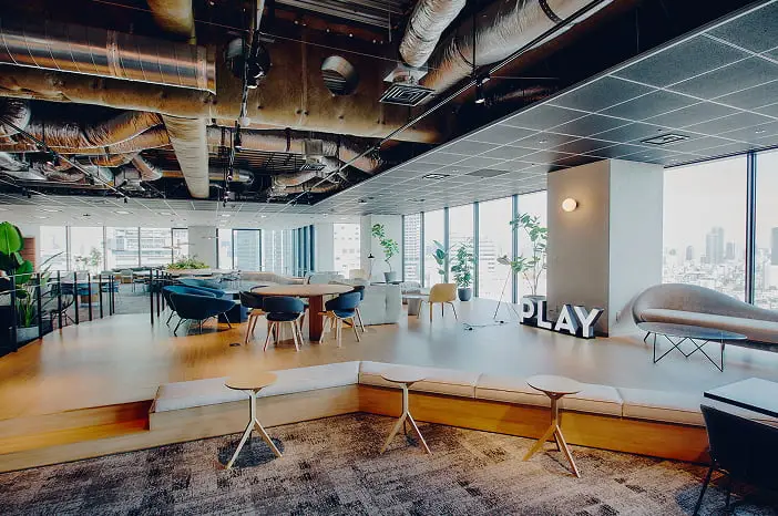 大きな窓から都市のスカイラインを望む、露出したダクトの天井と「PLAY」のサインが特徴的なモダンなオフィスラウンジ