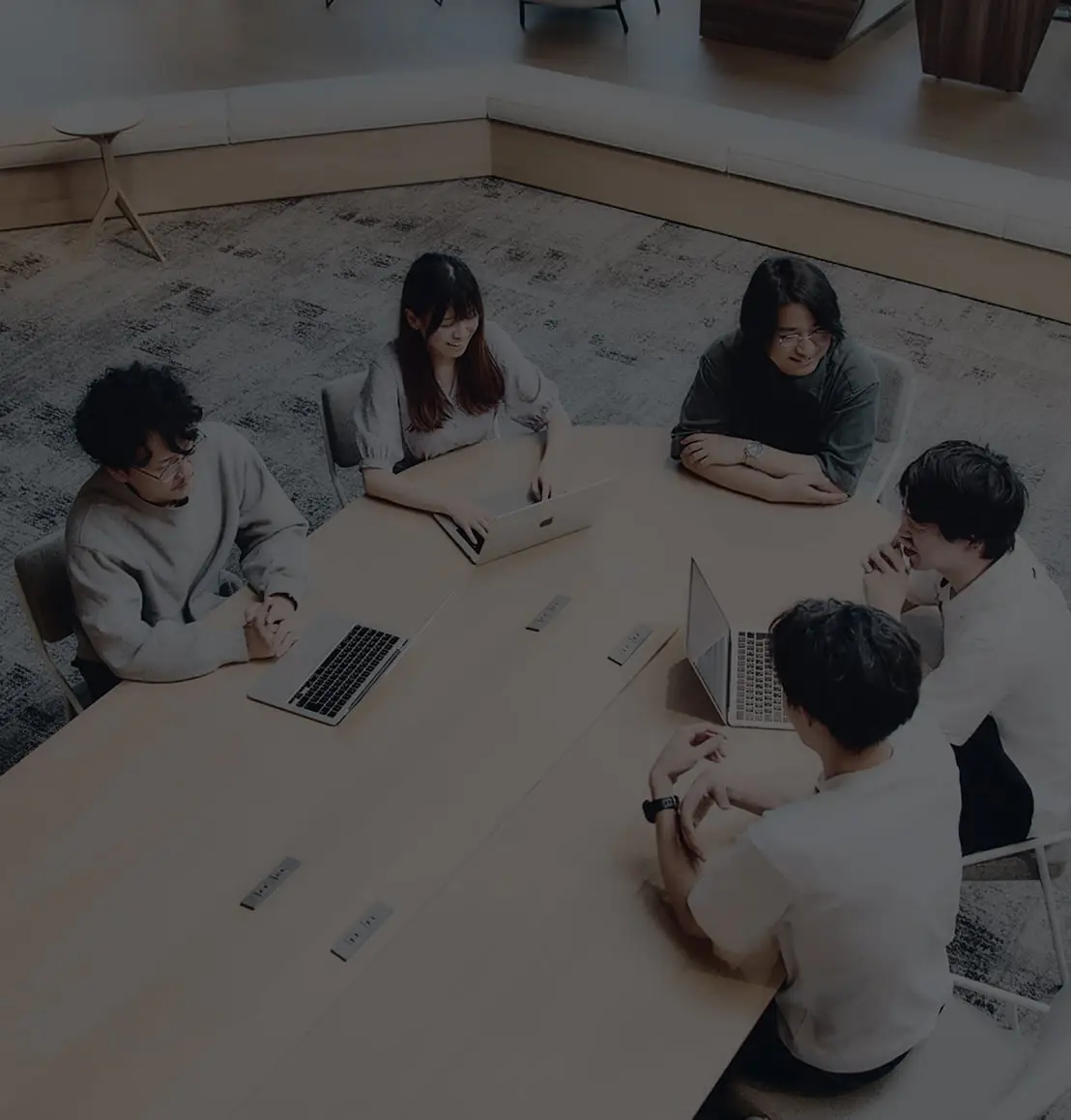 モダンなオフィスで、大きなテーブルを囲んでノートパソコンを使いながら共同作業や会話をする5人のビジネスパーソン