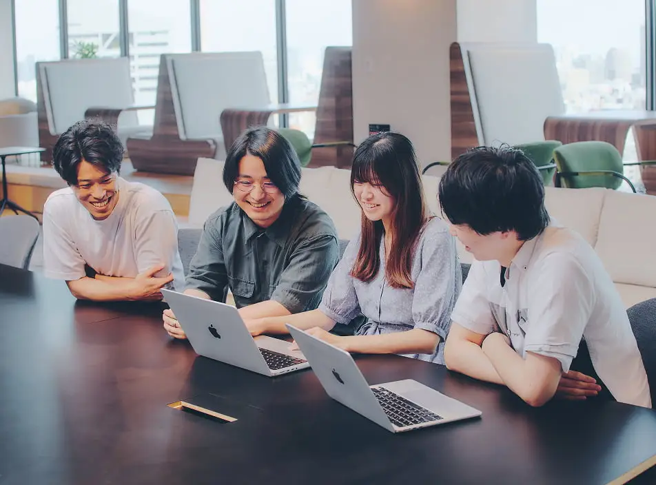 モダンなオフィスラウンジで、ノートパソコンを囲んで笑顔で共同作業をする4人の若者
