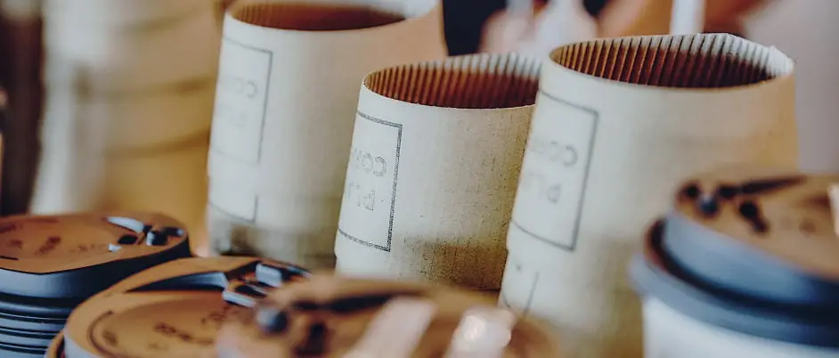 「PLAY CORP」のロゴが入った段ボール製のコーヒーカップスリーブと、積み重ねられた黒いカップの蓋が写るクローズアップ