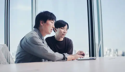 大きな窓から都市のスカイラインを望むモダンなオフィスで、2人の男性がノートパソコンの画面を指差しながら共同作業をしている様子