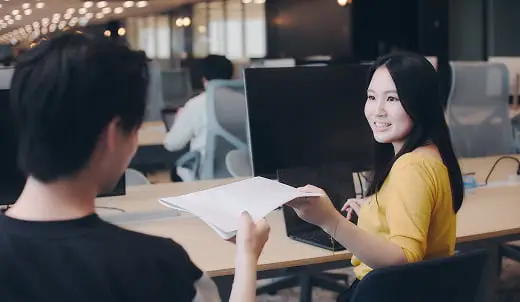 モダンなオフィスで、笑顔の若い女性が同僚から書類を受け取っている様子
