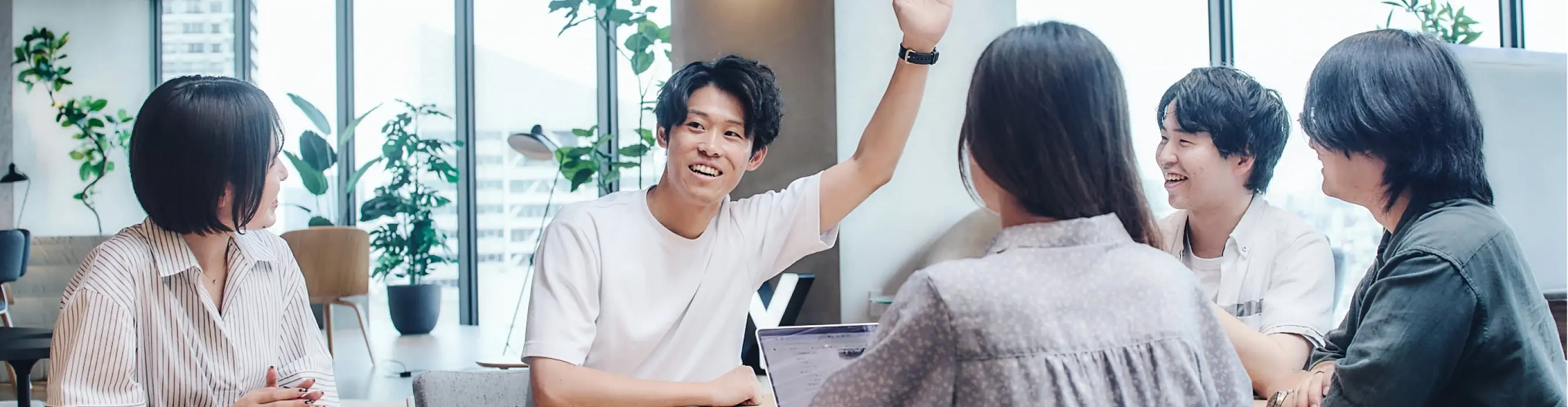 大きな窓から都市のスカイラインを望むモダンなオフィスで、会議テーブルを囲む5人の若いビジネスパーソンが活発に話し合い、一人が手を挙げている様子