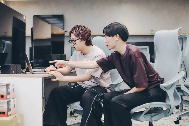 モダンなオフィスで、2人の若い男性がノートパソコンの画面を指差しながら共同作業をしている様子