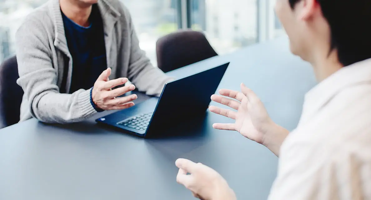 モダンな会議室で、ノートパソコンを挟んで2人のビジネスパーソンがジェスチャーを交えながら話し合っている様子