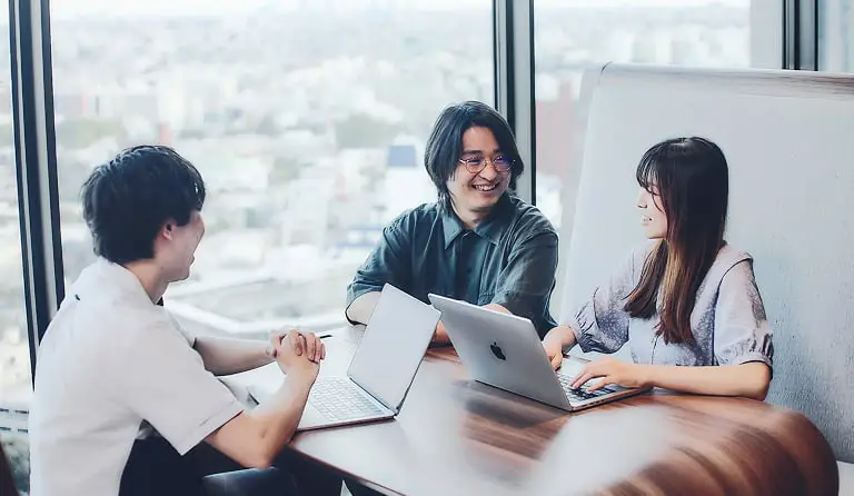 モダンなオフィスで、3人のビジネスパーソンがノートパソコンを使いながら笑顔で話し合っている様子