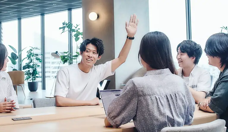 明るくモダンなオフィス空間で、5人の若いビジネスパーソンが会議テーブルを囲んで活発に話し合っている様子