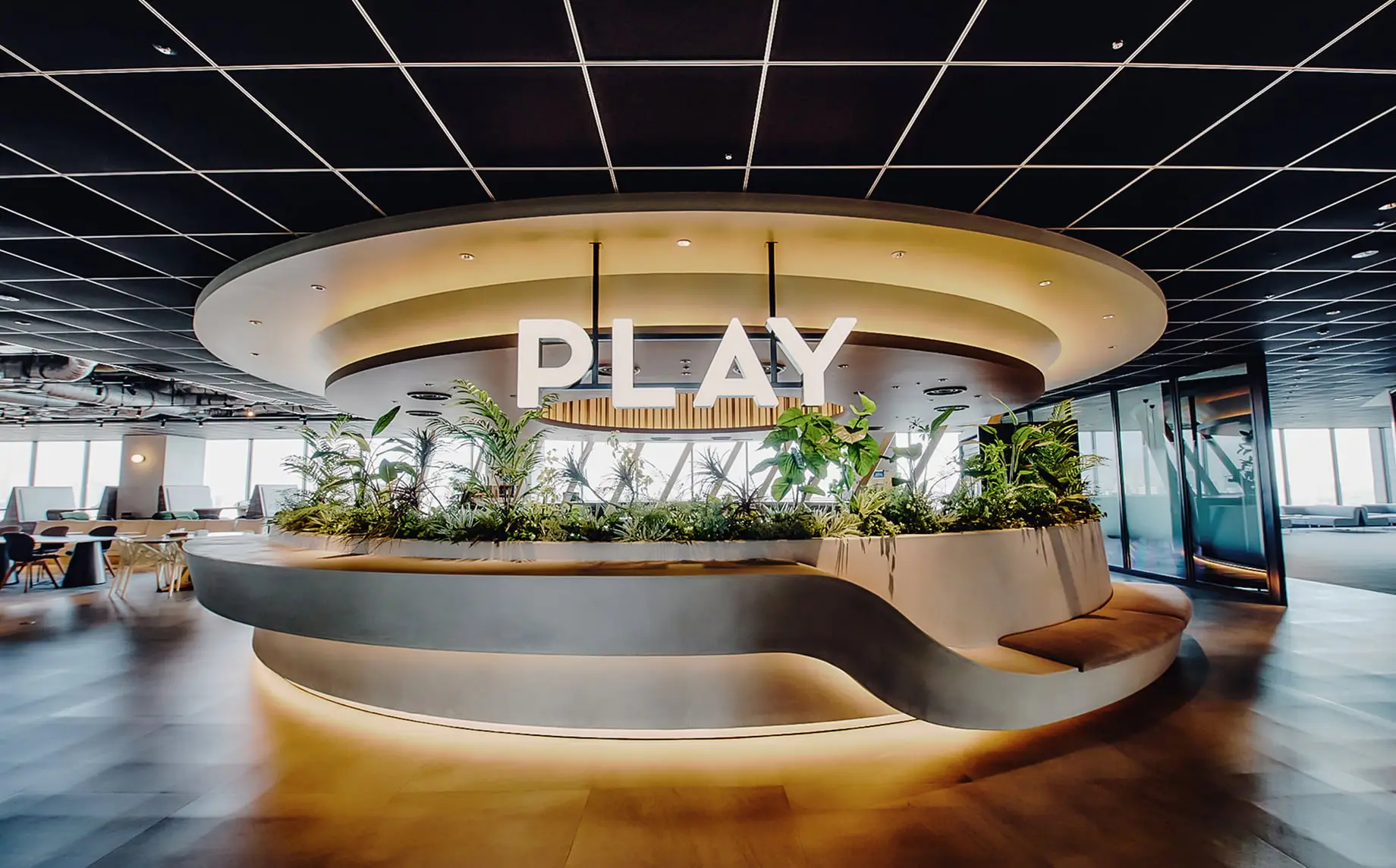 「PLAY」の文字が飾られた、植物が豊かな円形のプランターとモダンな座席があるオフィスラウンジ