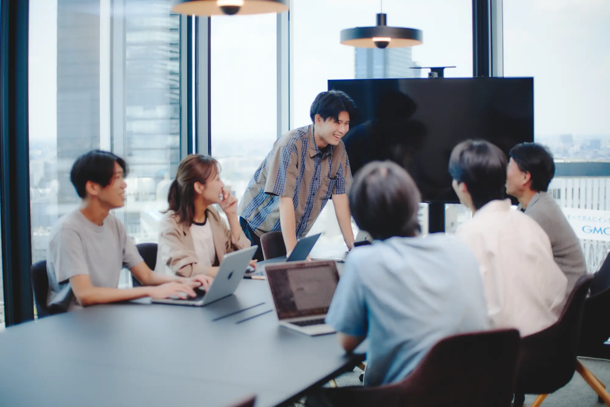 大きな窓から都市のスカイラインを望むモダンなオフィスで、会議テーブルを囲んでノートパソコンを使いながら笑顔で活発に話し合う6人の若いビジネスパーソン