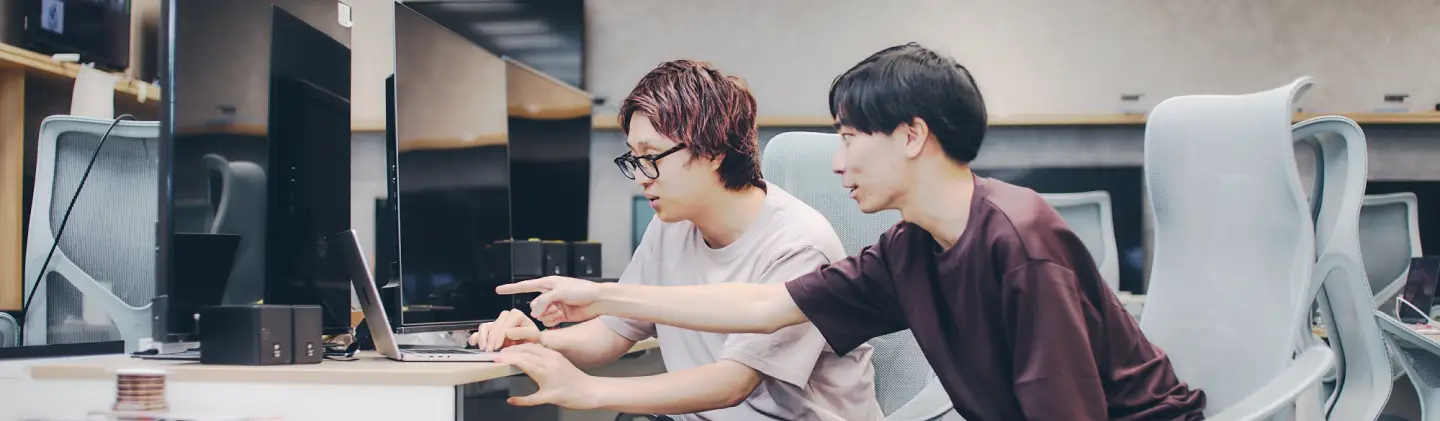 オフィスで2人の男性が並んで座り、ノートパソコンの画面を見ながら作業している。1人が操作し、もう1人が指を差して説明している様子。