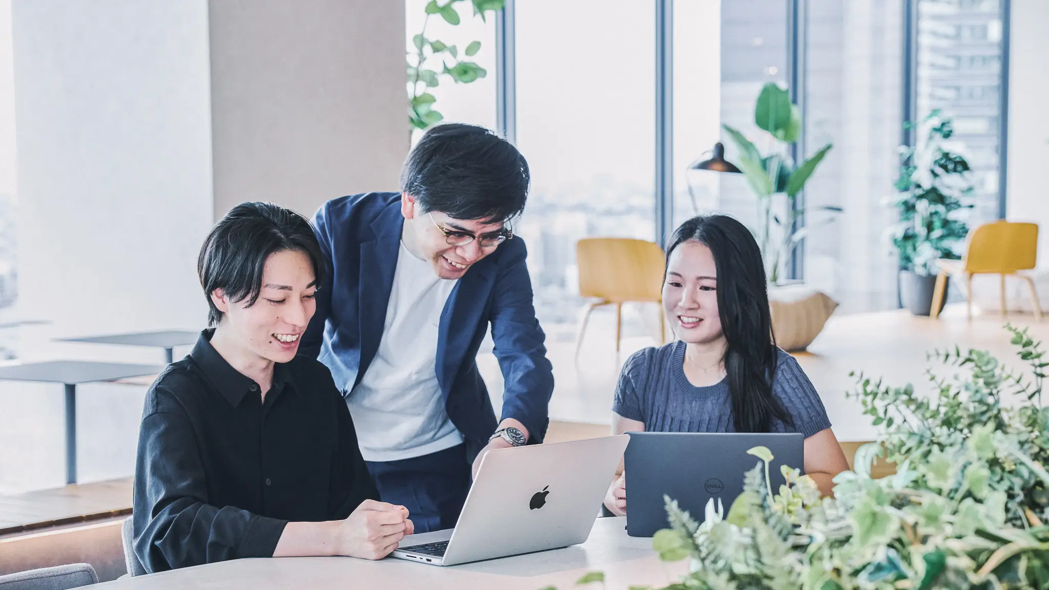 大きな窓から都市のスカイラインを望む明るいオフィスで、3人の若いビジネスパーソンが木製テーブルを囲み、ノートパソコンを使いながら笑顔で会話している様子