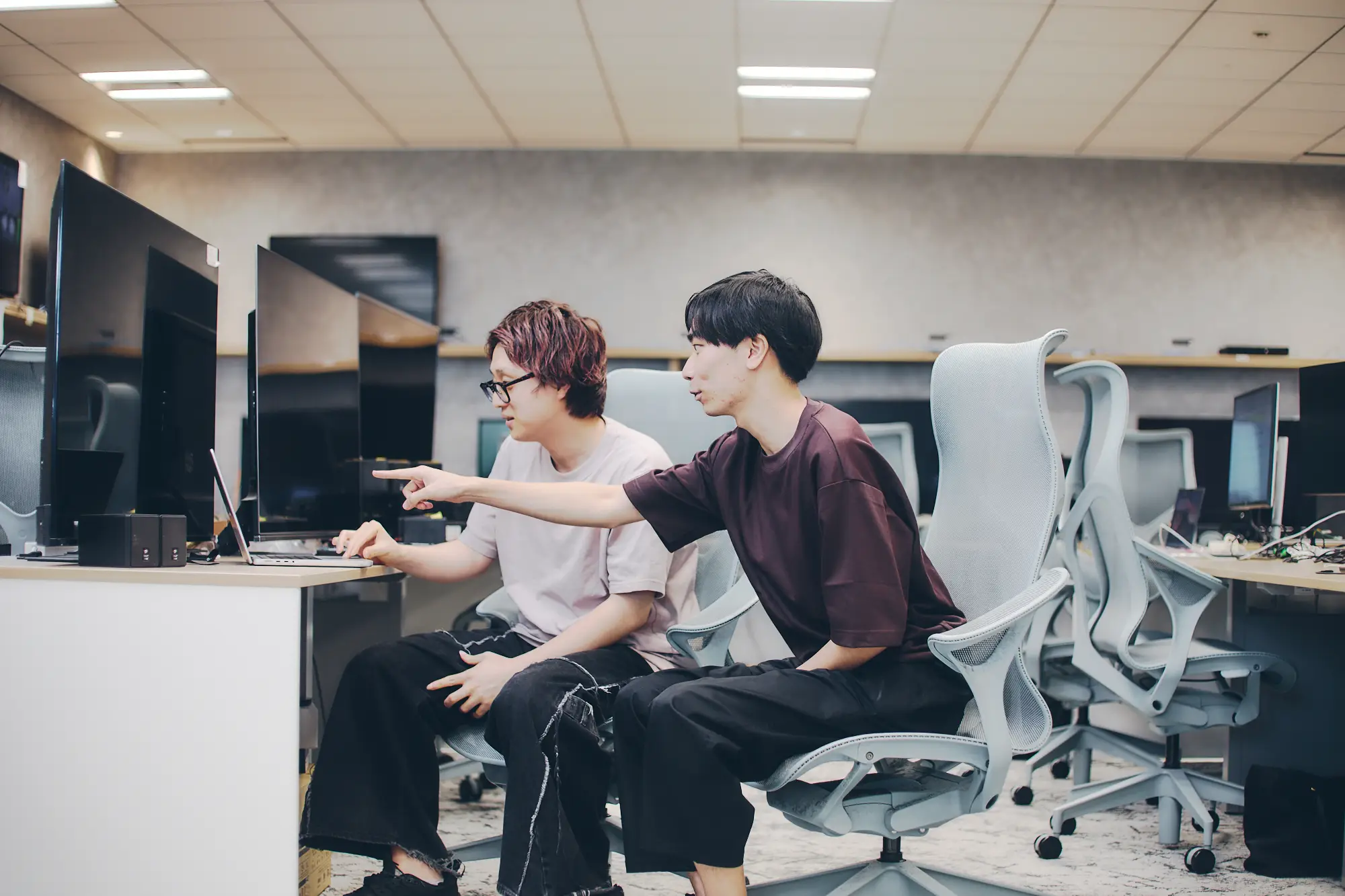 モダンなオフィスで、2人の若い男性がノートパソコンの画面を指差しながら共同作業をしている様子