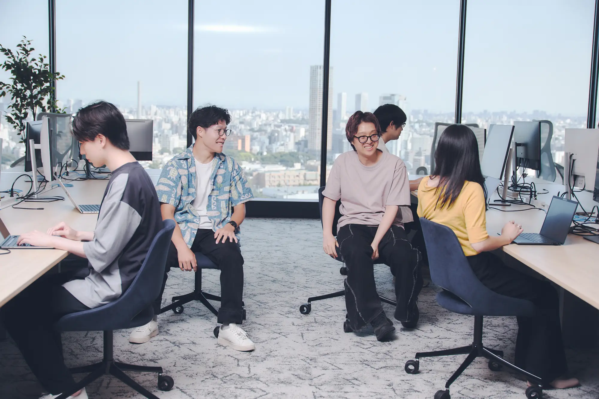 大きな窓から都市のスカイラインを望むモダンなオフィスで、4人の若いビジネスパーソンがノートパソコンを使いながら笑顔で共同作業や会話をしている様子