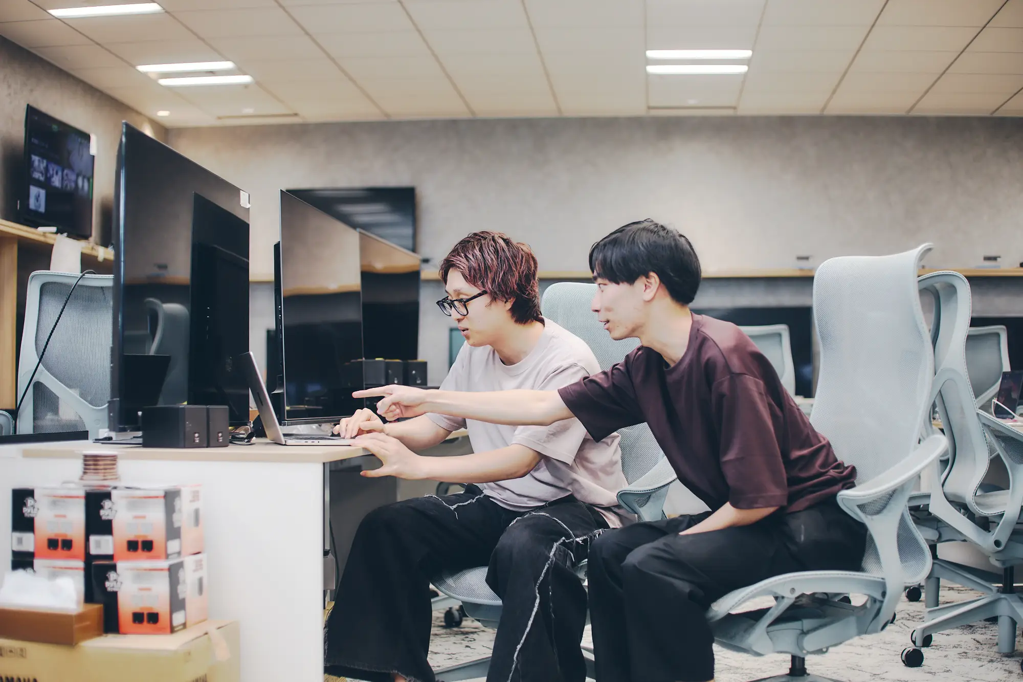 モダンなオフィスで、2人の若い男性がノートパソコンの画面を指差しながら共同作業をしている様子